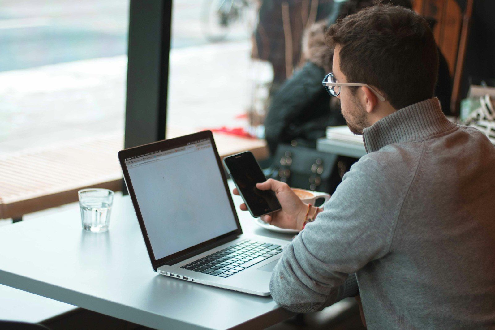 A man at his laptop checking his phone by a window
